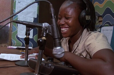 A smiling African woman wearing headphones and facing a microphone in a radio station.