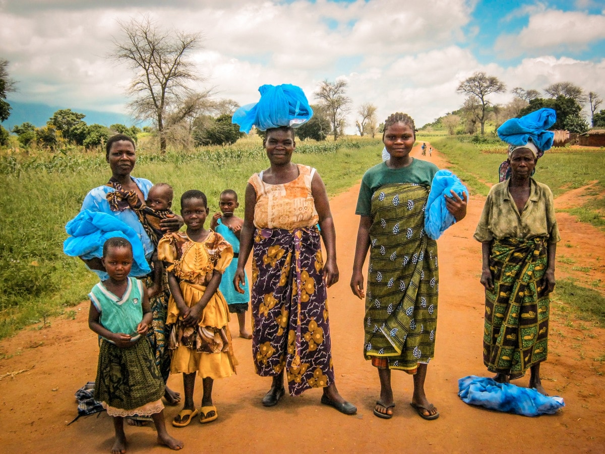 Four African women and four African children pose on a dirt road. Each of the women is carrying malaria nets.