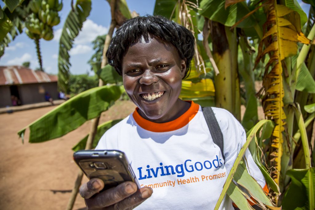 An African woman wearing a shirt that says "Living Goods: Community Health Promoter" looks up from phone and smiles.