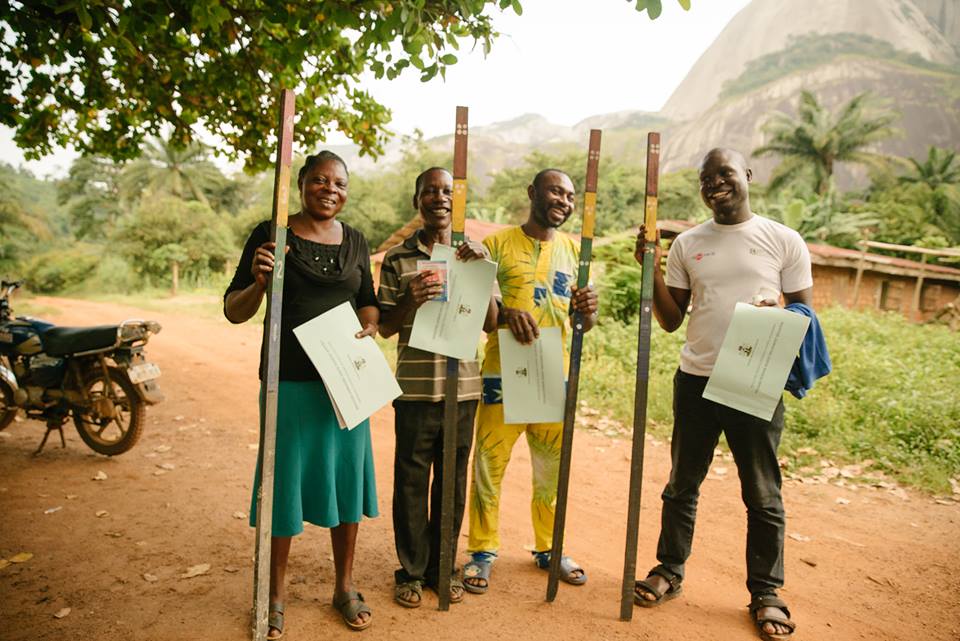Four Africans smiling, holding certificates and measuring sticks on a dirt road.